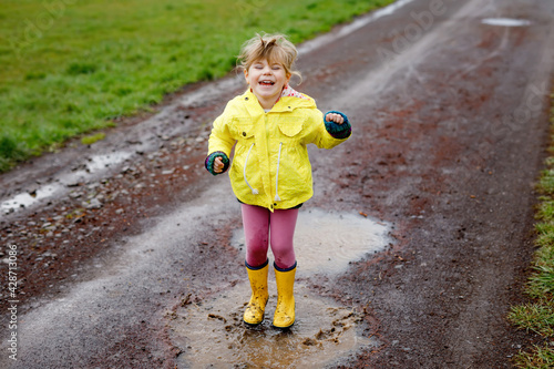 Little toddler girl wearing yellow rain gum boots, running and walking during sleet. Happy child in colorful clothes jumping into puddle, splashing with water, outdoor activity. Happiness, childhood