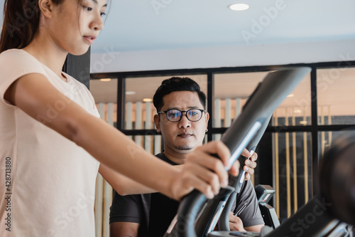 Asian man trainer checking program for help  support young customer woman exercise on elliptical cross machine in fit sportswear. Workout training in gym fitness for strong health  recreation concept.