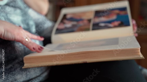 Elderly woman looking at old photo album close up. Adult female 60s holding nostalgic memory in photos in cozy modern home. Casual elderly life and wrinkled skin of hands. Biography or history concept