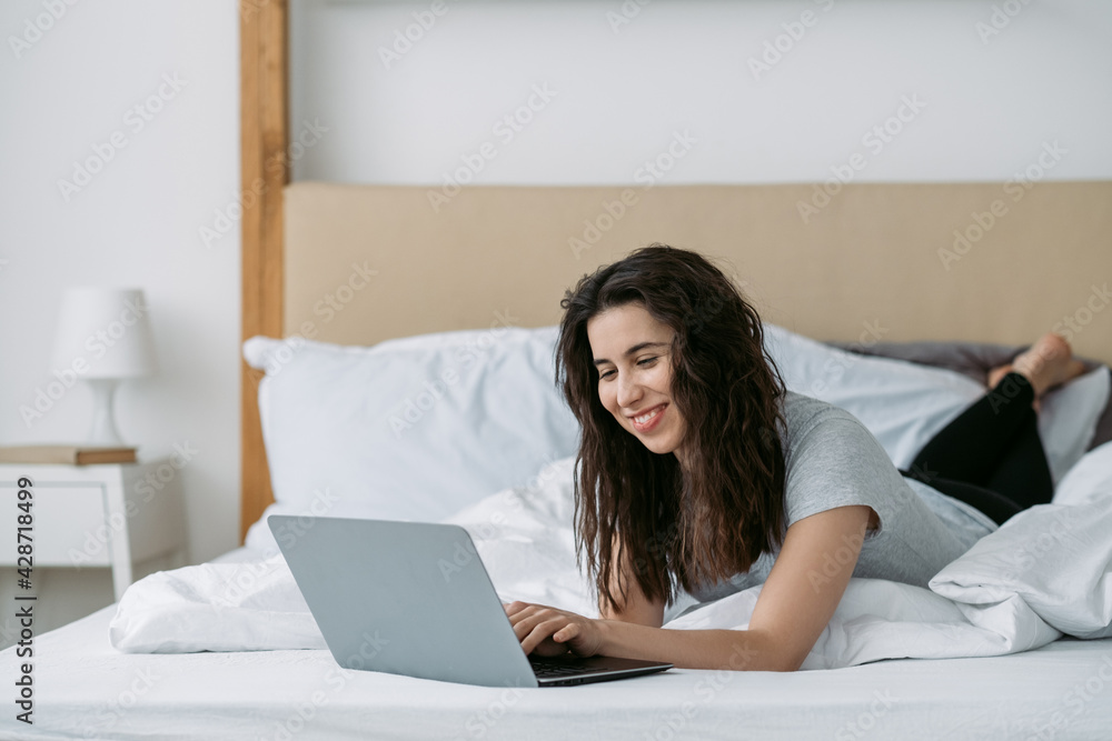 Morning video call. Online communication. Virtual meeting. Home leisure. Cheerful relaxed woman smiling using laptop lying on bed in light home bedroom. Morning video call. Online communication. Virtual meeting. Home leisure. Cheerful relaxed woman smiling using laptop lying on bed in light home bedroom.
