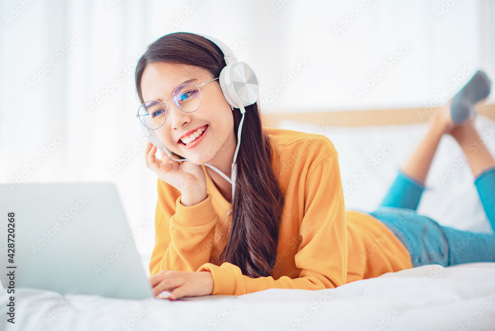 Happy woman relaxing while wearing headphones watching videos on laptop lying on a bed at home, Chill out and leisure concept Happy woman relaxing while wearing headphones watching videos on laptop lying on a bed at home, Chill out and leisure concept