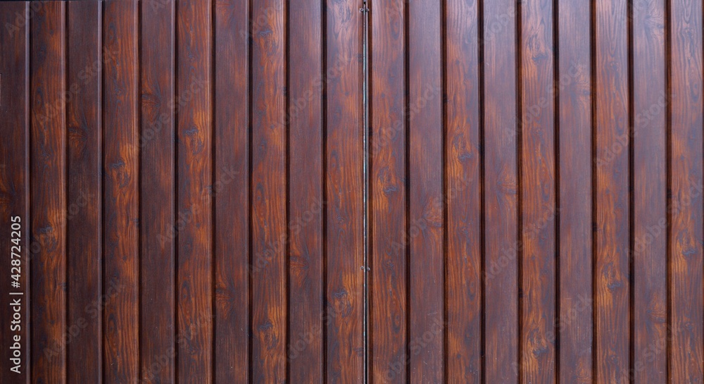 part of a fence or wall made of narrow metal panels stylized as dark stained wood, a fragment of gate leaves made of textured fake wood and with a flat smooth surface part of a fence or wall made of narrow metal panels stylized as dark stained wood, a fragment of gate leaves made of textured fake wood and with a flat smooth surface