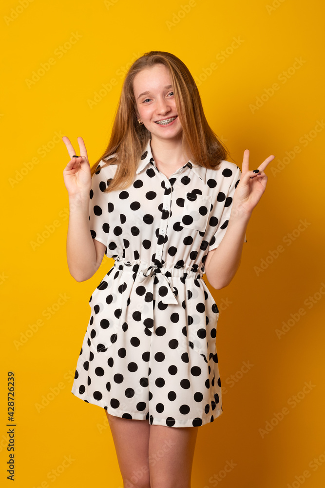 Cute little girl laughs and smiles on a yellow background. Happy teenage girl with braces on her teeth shows two fingers. Cute little girl laughs and smiles on a yellow background. Happy teenage girl with braces on her teeth shows two fingers.