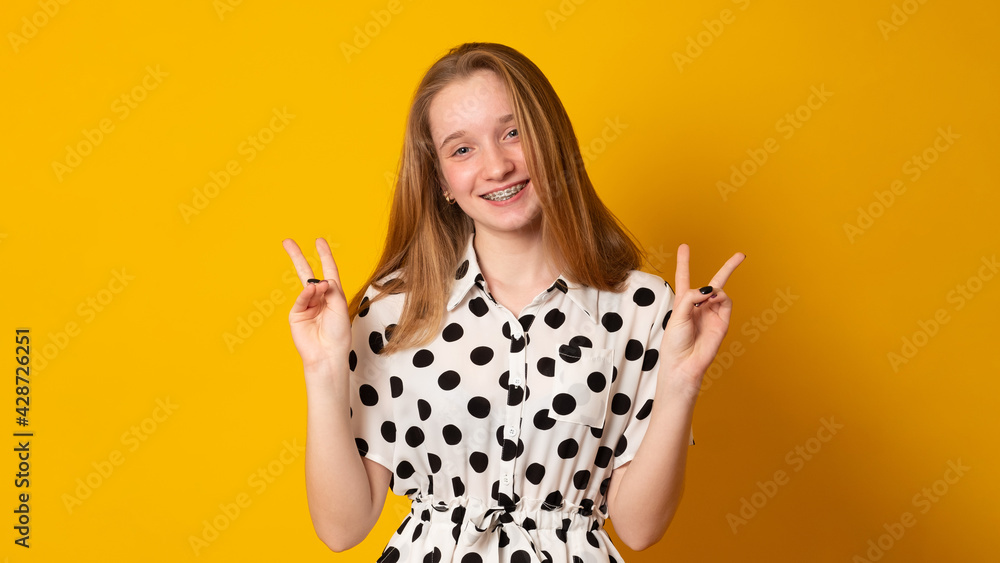 Cute little girl laughs and smiles on a yellow background. Happy teenage girl with braces on her teeth shows two fingers. Cute little girl laughs and smiles on a yellow background. Happy teenage girl with braces on her teeth shows two fingers.