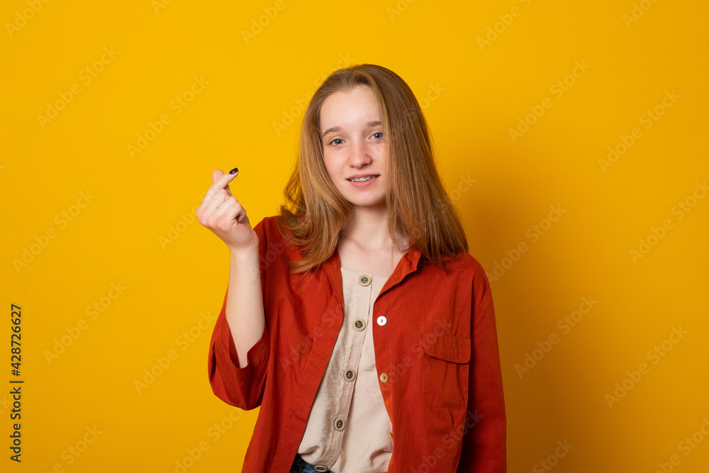 Cute girl with gentle smile makes korean like sign expresses love to you wears casual long shirt isolated over yellow background. Mini heart gesture. Body language concept. Cute girl with gentle smile makes korean like sign expresses love to you wears casual long shirt isolated over yellow background. Mini heart gesture. Body language concept.