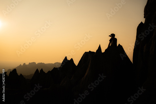 Silhouette of young Asian man sitting on beautiful sharp shape limestone mountains with one hand shading his eyes from the sun while admiring landscape in front of him with orange sky in background.