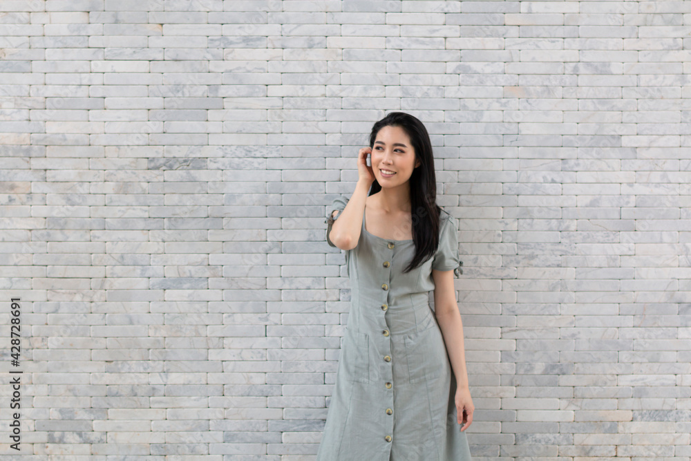 Portrait of a happy Asian girl in dress standing by the stone wall. Young traveling woman standing near stone wall during holiday. Tourist woman standing by the wall.
