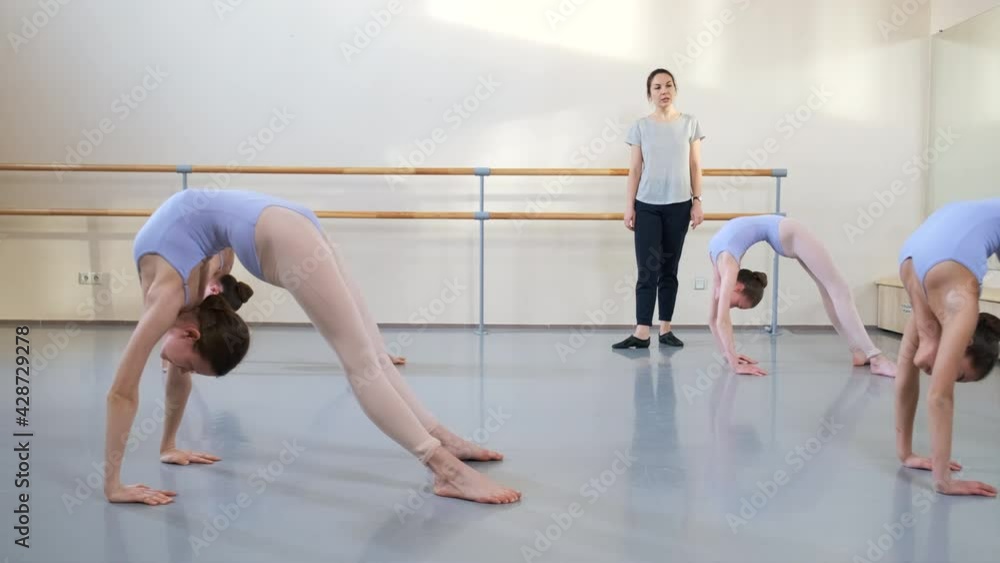Girls at ballet gymnastics class doing bridge pose exercise, dance school group. Their teacher trainer controls them. Flexible young gymnasts practicing wheel pose workout. Children in sport Girls at ballet gymnastics class doing bridge pose exercise, dance school group. Their teacher trainer controls them. Flexible young gymnasts practicing wheel pose workout. Children in sport