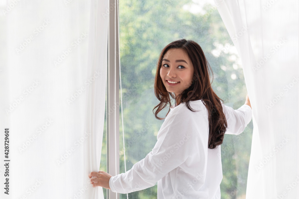 Young woman stand open white curtains at the window in morning after waking up in bedroom. Happy female opening window curtains at home. Woman standing by bedroom window opening curtains. Young woman stand open white curtains at the window in morning after waking up in bedroom. Happy female opening window curtains at home. Woman standing by bedroom window opening curtains.