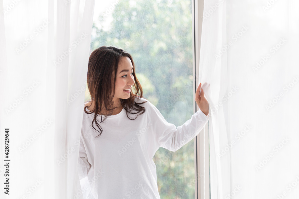 Young woman stand open white curtains at the window in morning after waking up in bedroom. Happy female opening window curtains at home. Woman standing by bedroom window opening curtains.