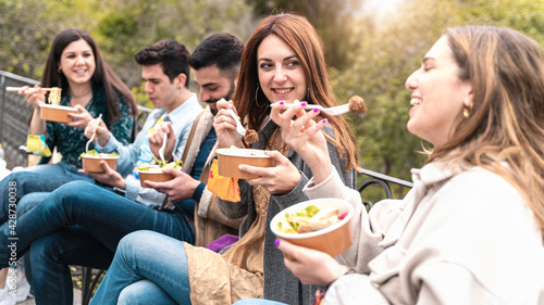 Gathering of young people eating take away organic food in plastic-free bowls and cutlery together in the park. Focus on the second from right smiling and talking.