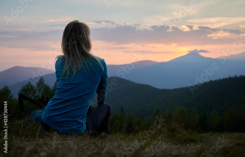 Back view of female hiker who resting after day of hike and watching at sunset above mountain beskids. Tourist enjoying landscape of mountain hills and evening sky when sun setting over horizon.