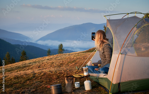 Rear view of girl photographing morning landscape with smartphone from her tent set up on hill during breakfast outdoors. Concept of active camping in the mountains, tourist equipment and photography. photo