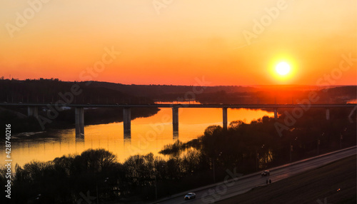 A crimson sunset on the background of a bridge over a wide river. © ru4eek