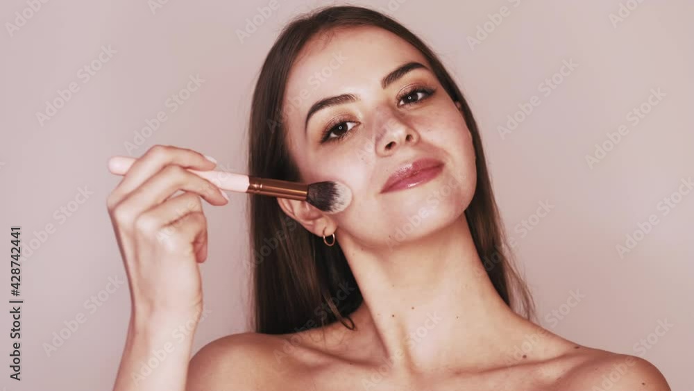 Nude makeup. Beauty care. Skin treatment. Portrait of smiling brunette woman with bare shoulders applying face powder with brush isolated on beige background. Nude makeup. Beauty care. Skin treatment. Portrait of smiling brunette woman with bare shoulders applying face powder with brush isolated on beige background.