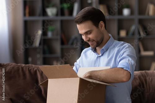 Excited millennial Caucasian man open unbox carton package shopping online on Internet from home. Happy young male unpack box buy purchases on web. Good delivery service, shipping concept.