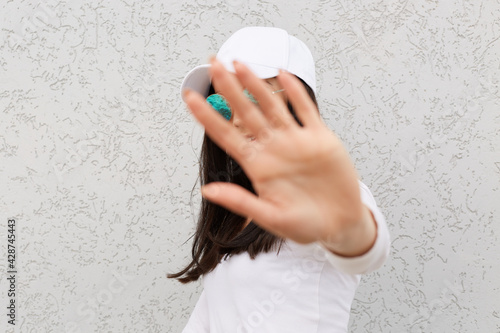 Positive dark haired female showing palm to camera, stop gesture, happy expression ,wearing white shier and baseball cap, shirt and sunglasses, posing isolated over light wall outdoor. photo