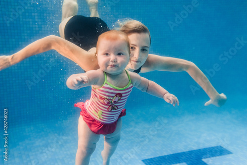 Mom holds daughter are immersed in water, swimming under water in paddling pool. Diving baby. Learning infant child to swim. Young mother or swimming instructor and happy little girl.
