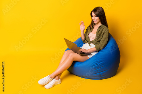 Photo of girl sit beanbag hold laptop wave hand screen wear green shirt skirt footwear isolated yellow color background