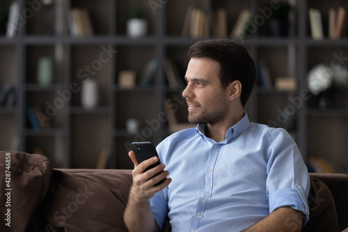Smiling young Caucasian man use cellphone gadget look in distance dreaming planning. Pensive millennial male browse wireless Internet on smartphone thinking or pondering. Vision, planner concept.
