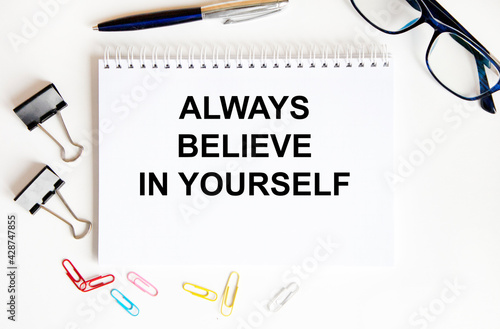 In the notebook text ALWAYS BELIEVE IN YOURSELF, next to glasses, paper clips, pen, paper clips.