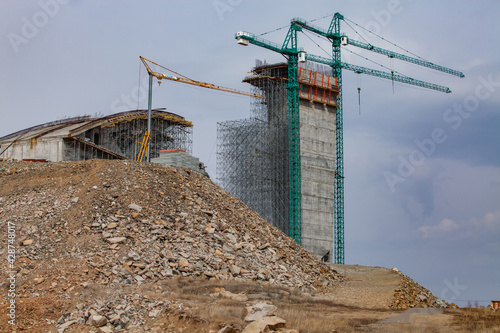 Construction of modern high-rise ski-jump in Shchuchinsk city, Kazakhstan. Cranes and concrete tower with scaffoldings. Heap of rocks on foreground photo