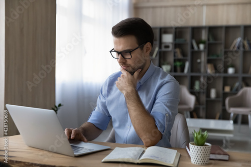 Pensive millennial male in glasses sit at desk at home office make notes work online on laptop gadget. Thoughtful young Caucasian man busy using computer device at workplace preparing report.