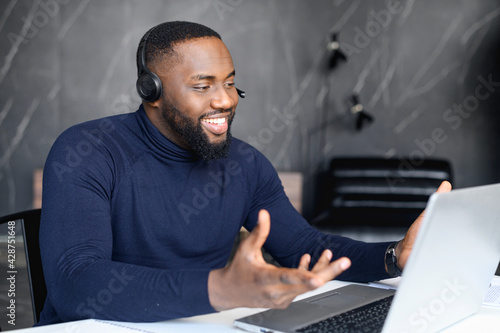 Focused young businessman in eyewear wearing headphones, holding video call with clients on laptop. Concentrated african man in glasses giving online educational class lecture, consulting customer