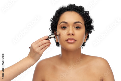 beauty, cosmetics and people concept - portrait of young african american woman and hand of make up artist with brush applying foundation to her face over white background