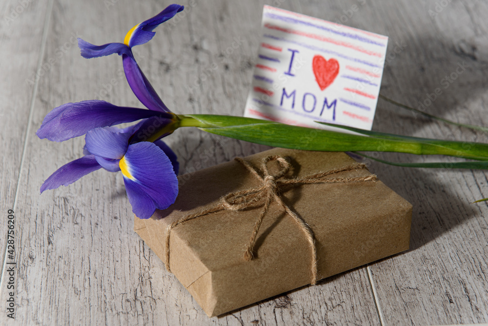 Gift box wrapped with kraft paper, card from child with message for mom and iris flower on gray wood table in morning sunlight. Mother's Day holiday concept.