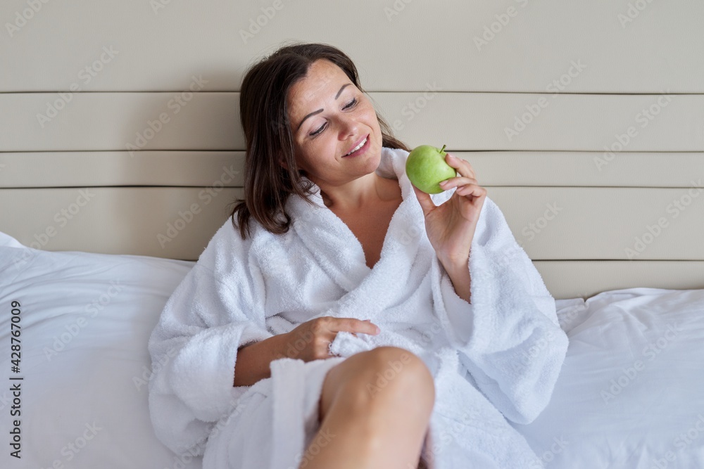 Middle-aged woman in white bathrobe with green apple in bed Middle-aged woman in white bathrobe with green apple in bed
