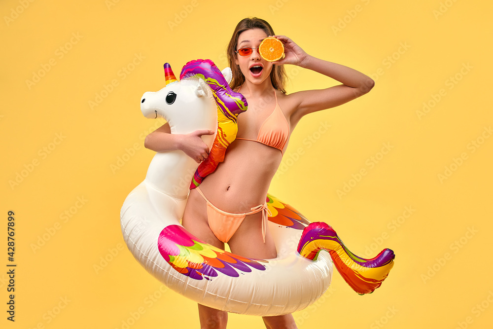 Cute woman in swimsuit with unicorn inflatable multicolored circle and orange slice isolated on yellow background. Summer time. Rest, vacations, travel.