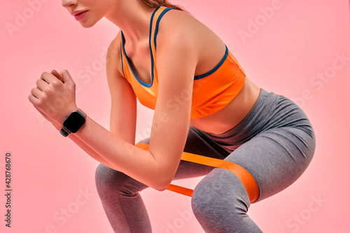 Cute sporty girl in sportswear doing exercises with sports elastic band isolated on pink background. Sports, yoga, active lifestyle, exercise for health.