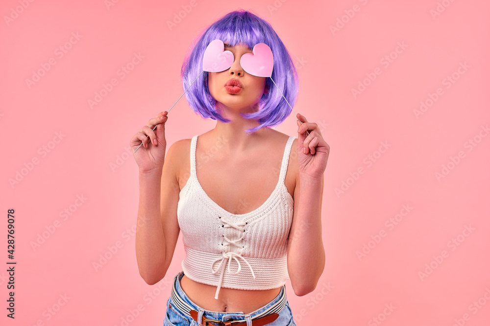 Beautiful young sexy girl in a purple wig is having fun holding valentine hearts on sticks isolated on a pink background.