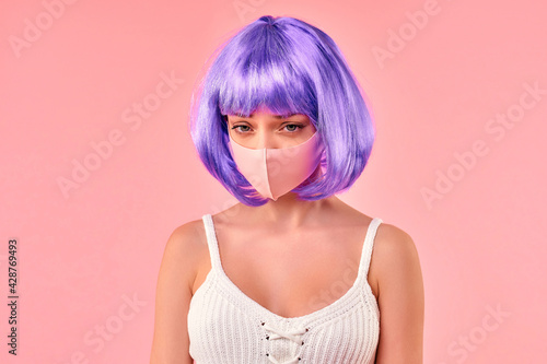 Beautiful young sexy girl in a purple wig in a protective medical mask isolated on a pink background.