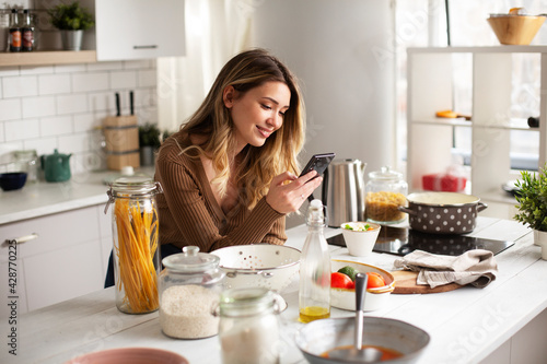 Young woman in kitchen. Beautiful woman using phone while making food..