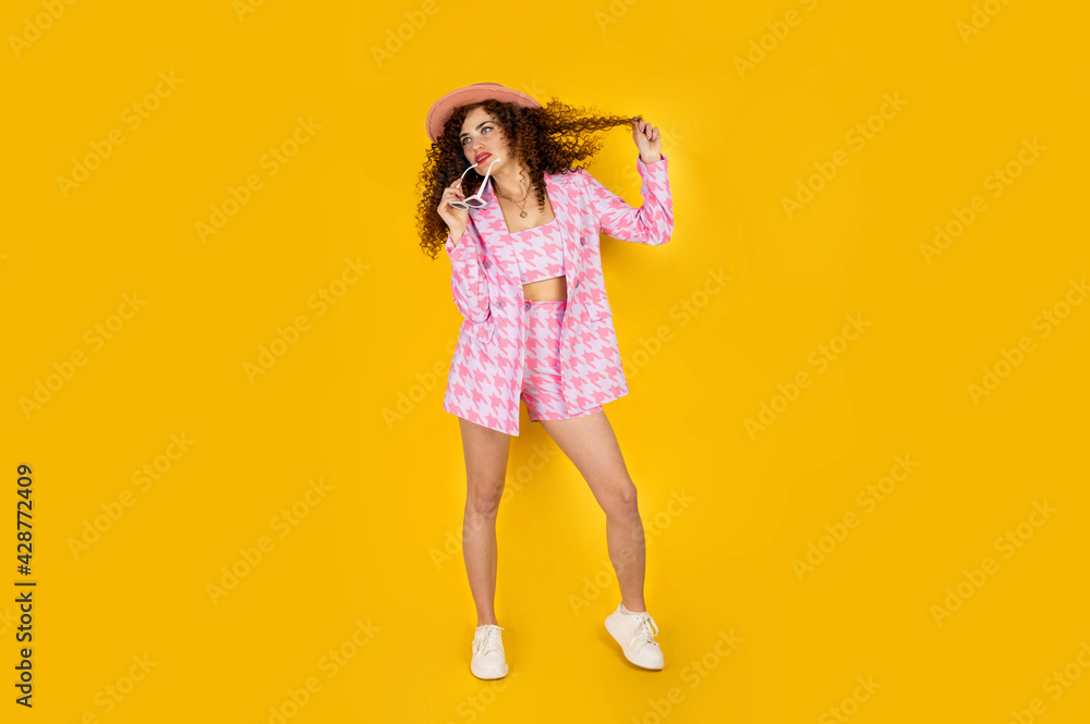 Girl with curly hair in summer pink outfit with hat smiling and enjoying. Posing on the yellow background. Girl with curly hair in summer pink outfit with hat smiling and enjoying. Posing on the yellow background.