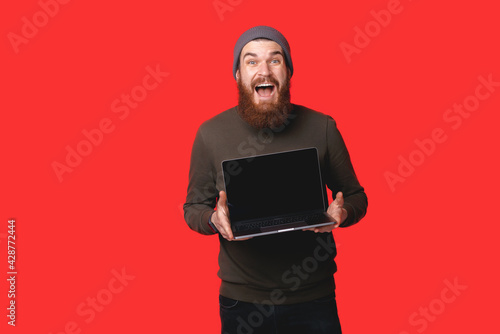 Amazed bearded hipster man showing laptop over red background