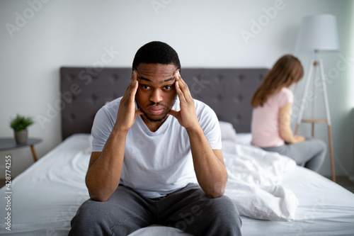 Sex or relationship difficulties. Unhappy black guy sitting on bed, massaging temples, overwhelmed with family problems photo