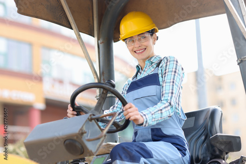 Young woman builder in hard hat sitting behind wheel of asphalt paver