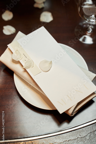 there is a white plate on the table on which there is a napkin and a menu and white rose petals