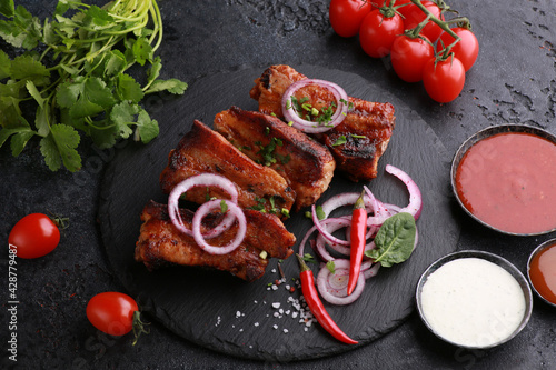 Meat and barbecue. Fried pork ribs with fresh herbs, cilantro, onions, peppers, spices and vegetables on a black table. Assorted sauces in a bowl. Background image, copy space