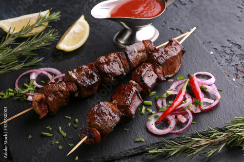 Fried liver shish kebab with lard on skewers with rosemary, lemon, fresh vegetables, herbs and tomato sauce on black table. Background image, copy space