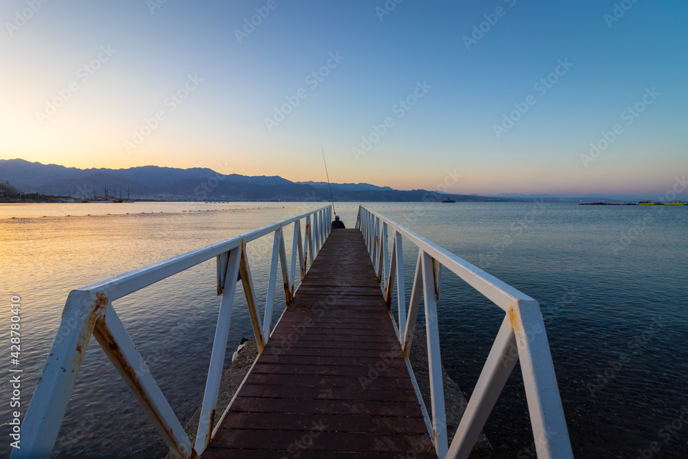 A wooden and metal path into the Red Sea, at the end sits a fisherman with a fishing rod, sunrise