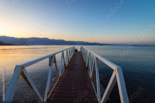 A wooden and metal path into the Red Sea  at the end sits a fisherman with a fishing rod  sunrise