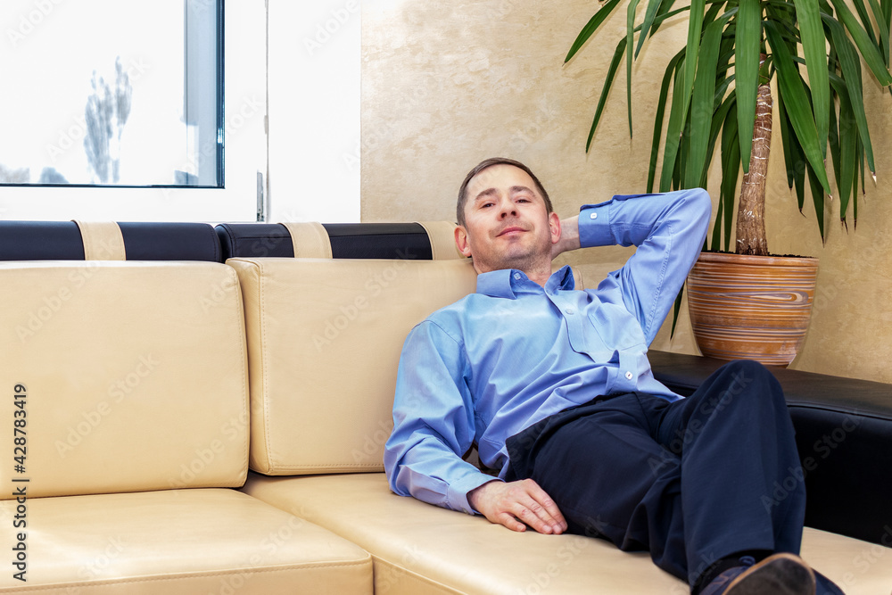 The concept of the need for rest during work. A tired manager in a blue shirt is lying on the couch in his modern office. The concept of the need for rest during work. A tired manager in a blue shirt is lying on the couch in his modern office.