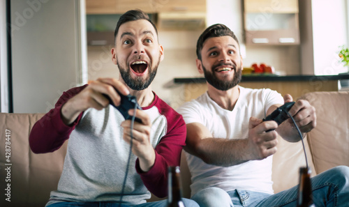 Excited smiling men playing in video games on tv at home on the couch. Friends with joysticks play game with happy emotions on faces photo