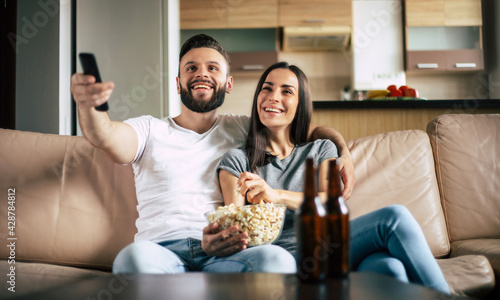 Beautiful smiling modern couple in love is watching some film or movie on TV while they relaxing on the couch at own apartment