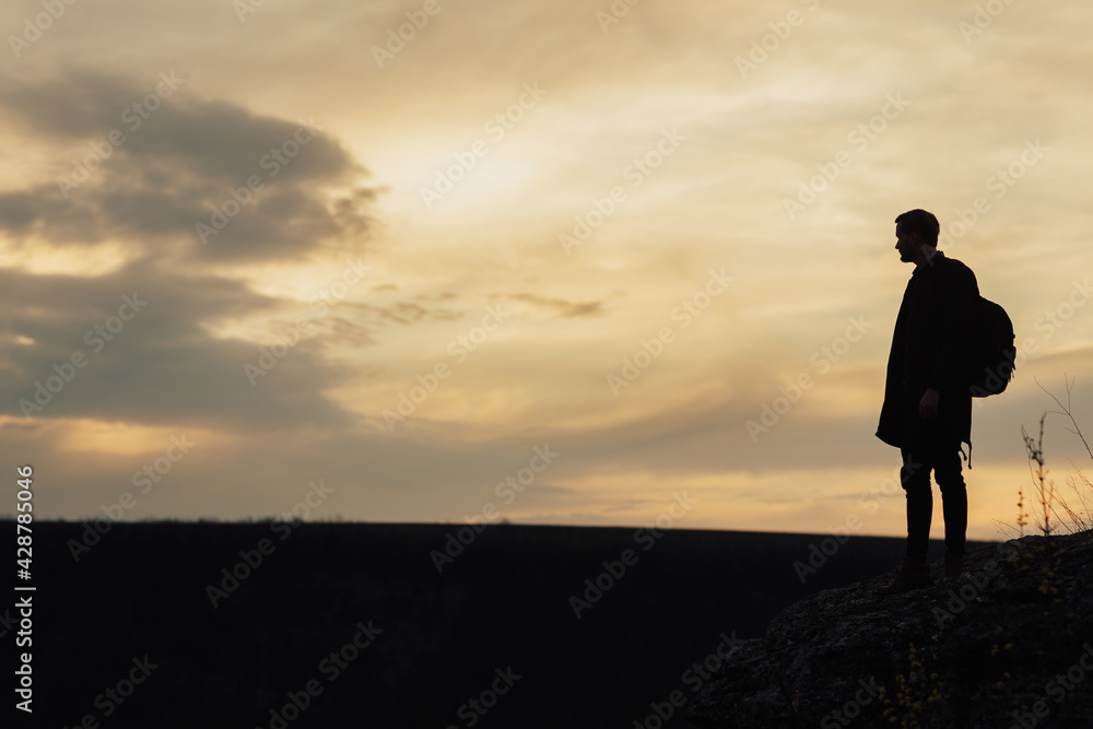 Dark silhouette of young man with backpack standing an evening sunset sky with copy space for your advertisement.