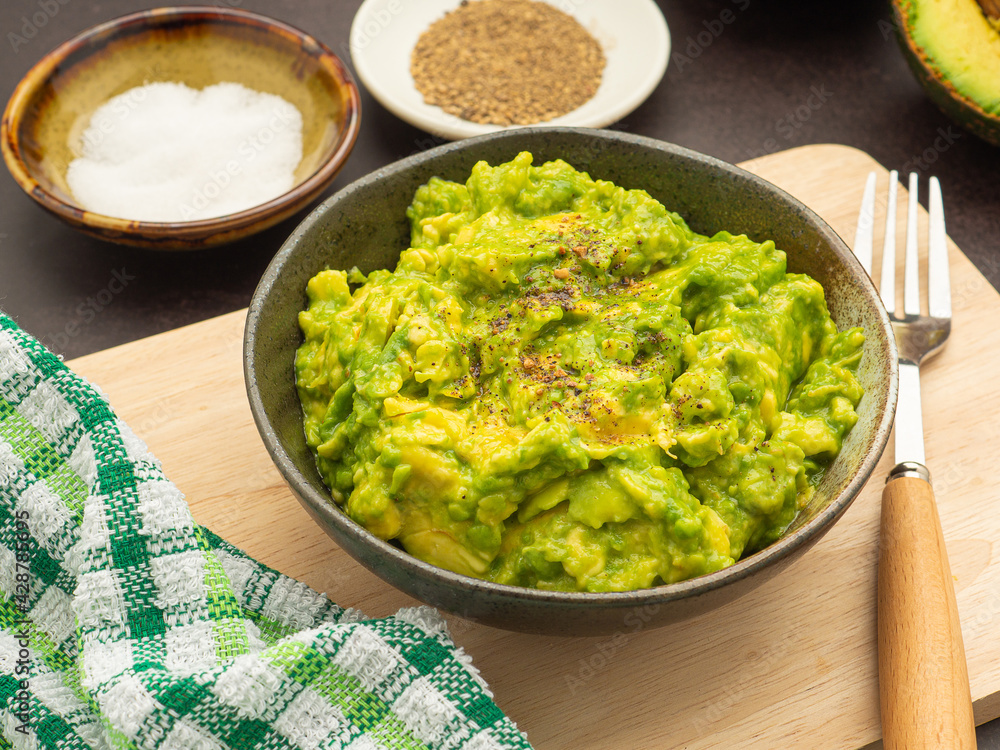 Fresh guacamole on a dish placed on a cutting wooden board with ingredients for homemade guacamole: avocados, lemon, salt, and pepper. Top view.  Concept of traditional Mexican preparation